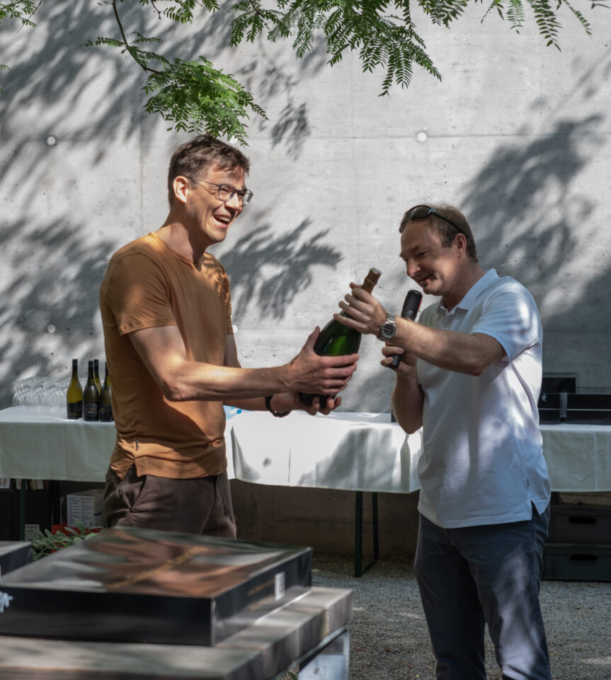 Two people stand outdoors under dappled shade. They hold a bottle of sparkling wine in their hands; a table with bottles and a concrete wall are in the background.