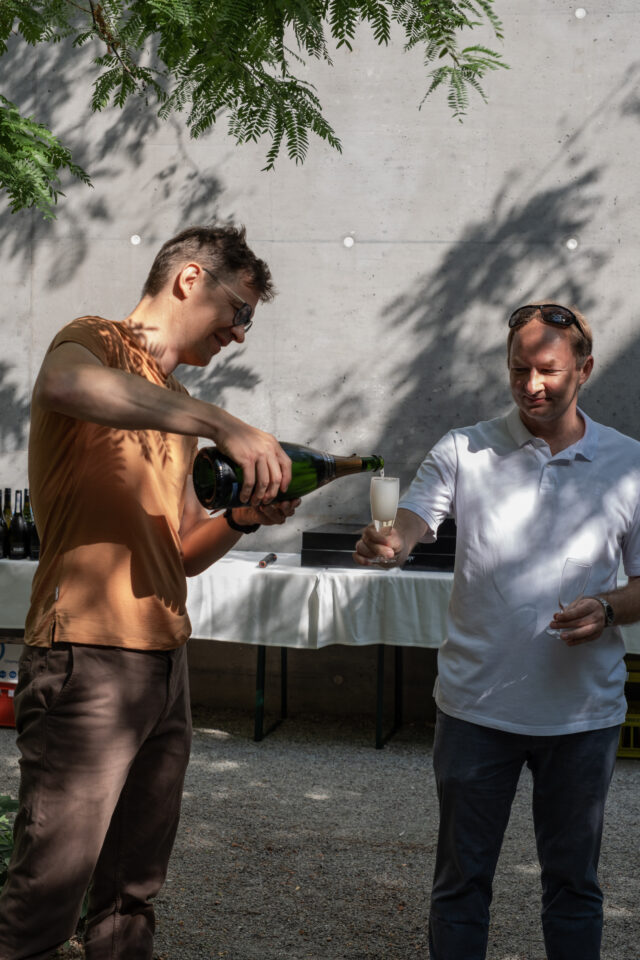 Two people stand outdoors under dappled shade. One pours sparkling wine from a green bottle into a flute held by the other, who smiles; a table with bottles and a concrete wall are in the background.
